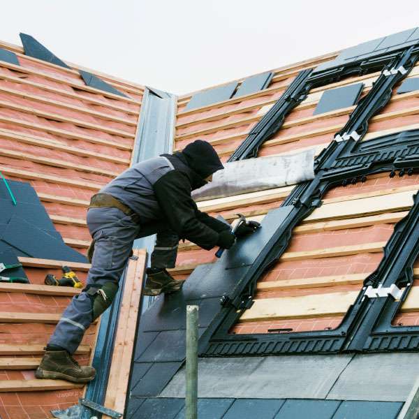 Roofer installing shingles and waterproofing materials around a skylight on a pitched residential roof under construction.