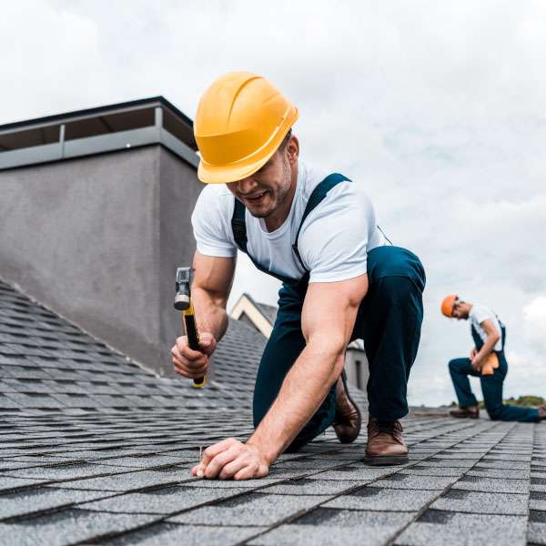 Roofing contractor using a hammer to secure shingles on a residential roof while another worker inspects in the background.